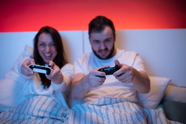 Couple of gamers in love passionately playing video games in\
bed with joysticks