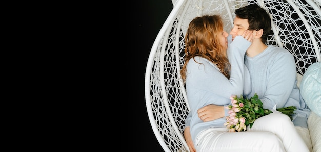 Couple girl and guy hugging on a big chair with a bouquet of roses. White and blue sweater.