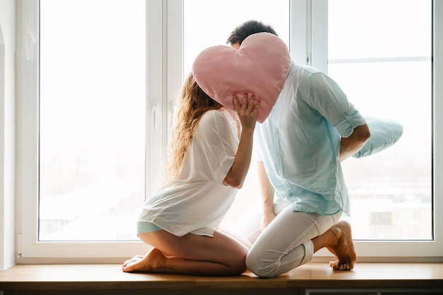 Couple girl and guy play with pillows near window. White and blue clothes. Valentine day.