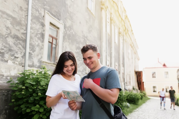 A couple of handsome heterosexual tourists in love looking at a city map to understand how to find a place. The girl points her finger and smiles. Wife and husband have a vacation trip to other city.