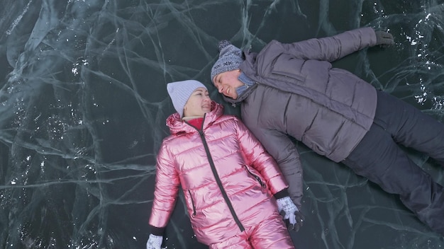 커플은 얼어붙은 호수의 얼음을 배경으로 겨울 산책을 즐깁니다. 연인들은 균열이 있는 맑은 얼음 위에 누워 재미있는 키스와 포옹을 합니다. 위에서 보기 눈 덮인 얼음 위의 행복한 사람들 허니문 러브 스토리