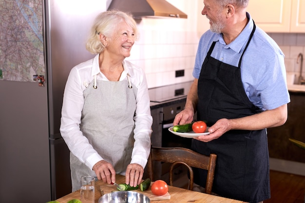 couple having fun in kitchen with healthy food, cooking meal at home, preparing lunch with bio fresh vegetables, carving or cutting vegetables, man helps his wife, wearing apron