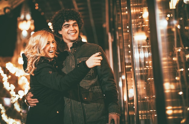 Couple having fun outdoors at Christmas time pointing at something behind shop window.