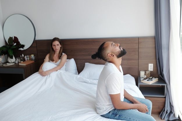 Couple having problems in bed, upset man sitting on bed, his unhappy wife sitting under blanket in background