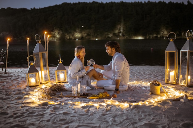 Coppia con una cena romantica, abbracciando e tintinnando bicchieri di vino nel luogo splendidamente decorato illuminato con luci diverse la spiaggia sabbiosa al tramonto