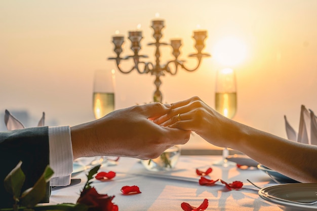 Пара, взявшись за руки, имея дату обеда в ресторане в вид на закат. Валентина, пара, медовый месяц, ужин, вино, романтическая концепция.