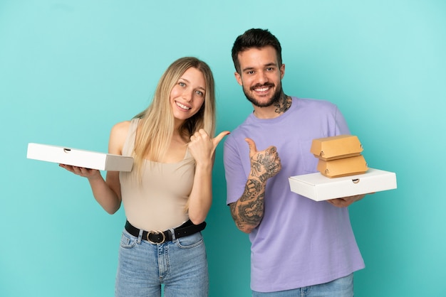 Couple holding pizzas and burgers over isolated blue background giving a thumbs up gesture with both hands and smiling