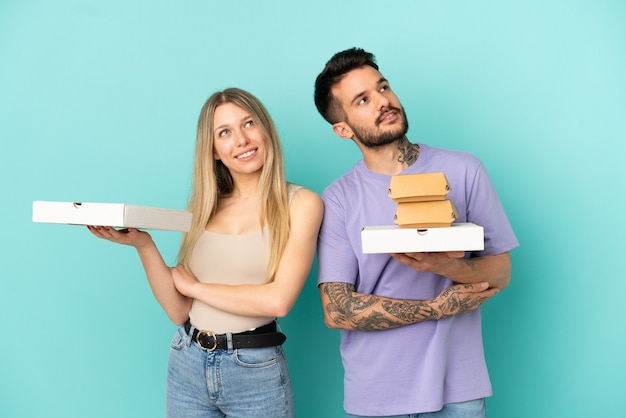 Couple holding pizzas and burgers over isolated blue background looking up while smiling