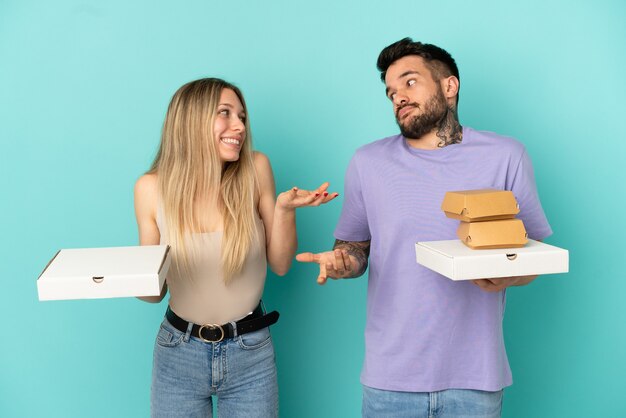 肩を持ち上げながら重要でないジェスチャーを作る孤立した青い背景の上にピザやハンバーガーを保持しているカップル