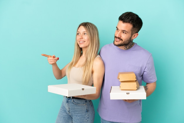 Пара, держащая пиццу и гамбургеры на изолированном синем фоне, представляя идею, глядя в сторону