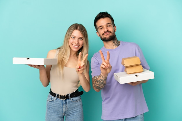 Couple holding pizzas and burgers over isolated blue background smiling and showing victory sign