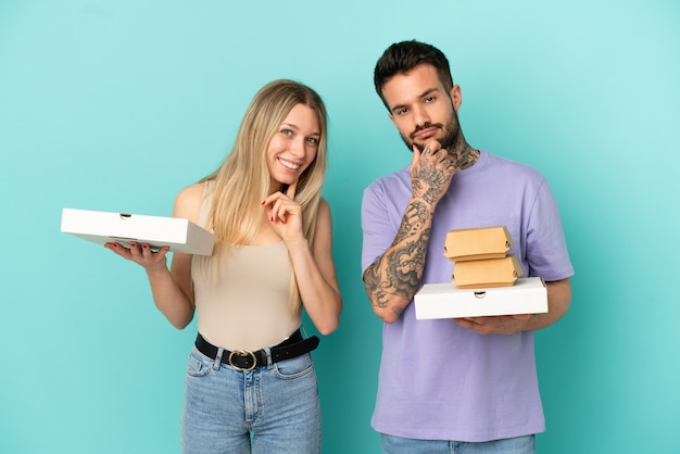 Пара, держащая пиццу и гамбургеры на изолированном синем фоне, улыбаясь со сладким выражением лица