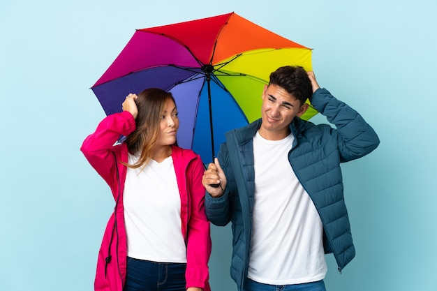 couple holding an umbrella on blue having doubts while scratching head