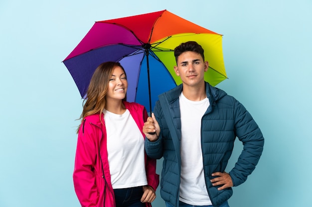 Couple holding an umbrella on blue posing with arms at hip and smiling