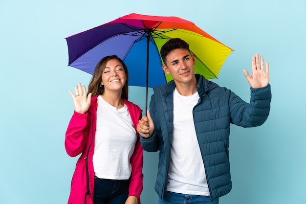 Couple holding an umbrella on blue saluting with hand with happy expression