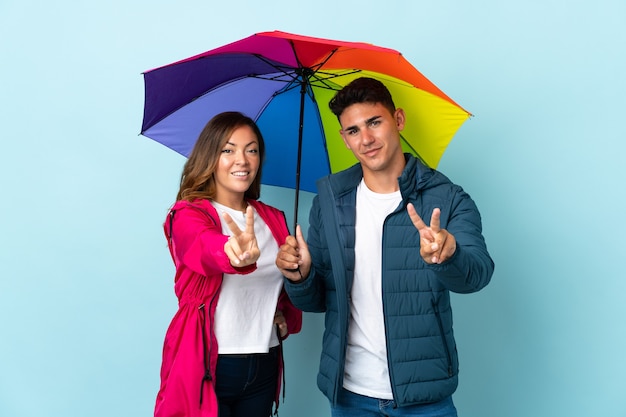 Couple holding an umbrella on blue smiling and showing victory sign