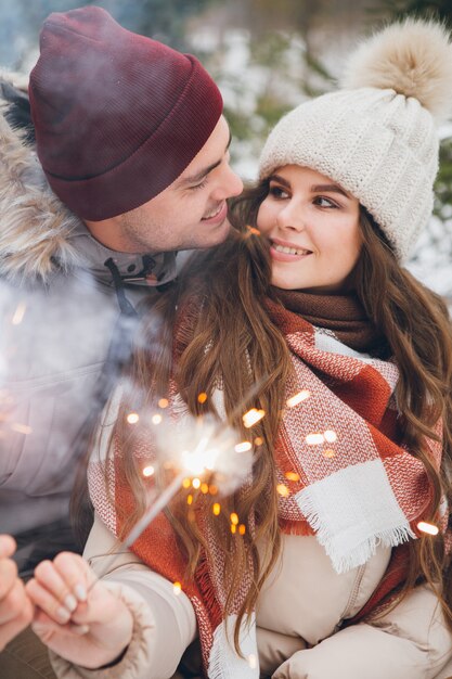 Couple hugs and burns sparklers in a winter coniferous forest while sitting on a sled