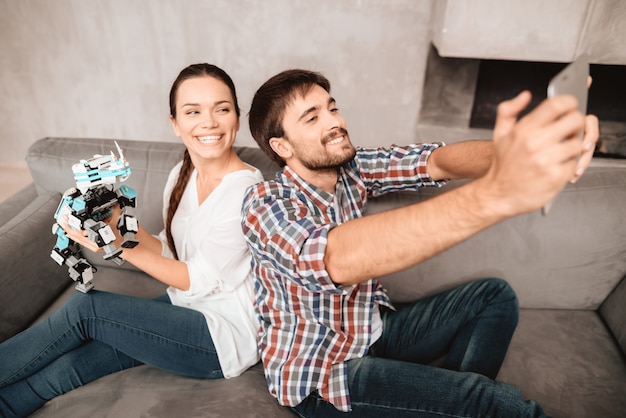 Couple is sitting on the couch and doing selfie with robot.