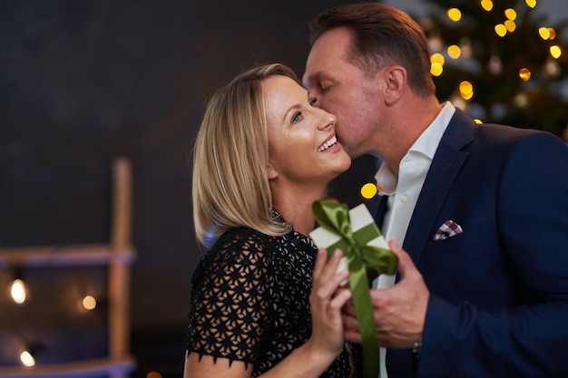 Couple kiss in front of chrismas tree holding gift. High quality photo