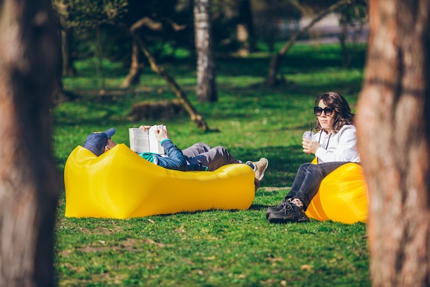 Couple laying on yellow inflatable mattress in city park reading book drinking smoothies