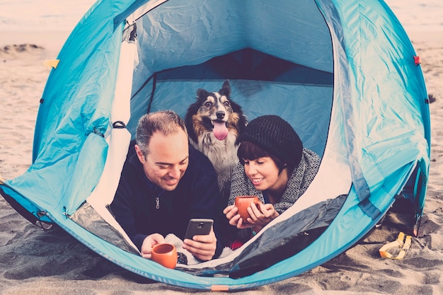 スマートフォンを見ているカップルとビーチでの無料キャンプでテントの中で楽しんでくださいカメラを見ている彼らの後ろの犬のボーダーコリー。ヴィンテージの色と休暇の家族の概念。代替t