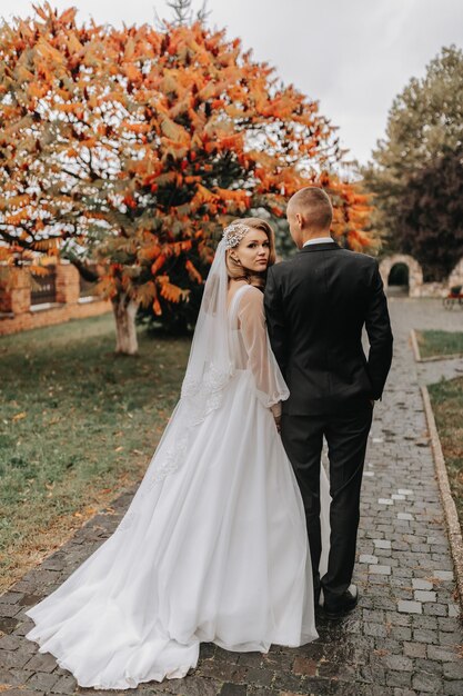 秋の公園で恋愛するカップル 袖の花束と花束の花束の結婚式のドレスを着た金の花嫁 クラシックな黒いスーツと白いシャツとネクタイを着た新郎