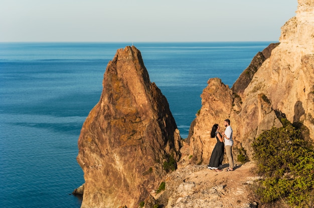 崖の端で抱き締める海沿いの愛のカップル。男は女の子に提案します。山での新婚旅行。男と女の旅行。結婚式。旅。愛。海で休んでいる新婚夫婦