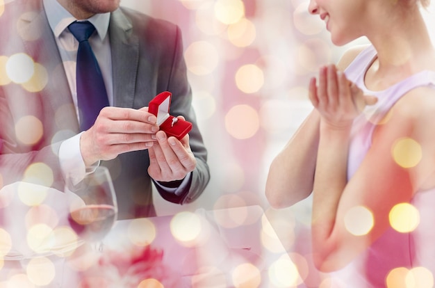 カップル、愛、婚約、休日のコンセプト – 興奮した若い女性と彼氏が、ピンク色の休日ライトの背景にレストランで指輪を渡す様子をクローズアップ