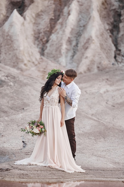 Couple in love in fabulous mountains hugging, Martian landscape. Lovers walk in the mountains in the summer, the girl in long light summer dress with a bouquet of flowers and a wreath on her head