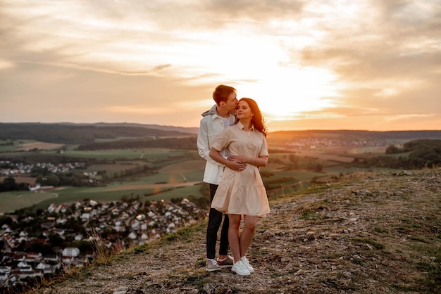 A couple in love, a guy and a girl are standing on a mountain at sunset hugging and kissing