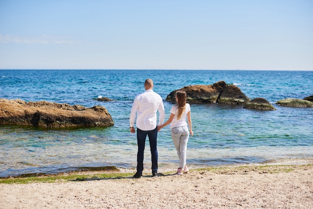 Couple in love hold hands on a tropical beach with a turquoise water and rocks in the background