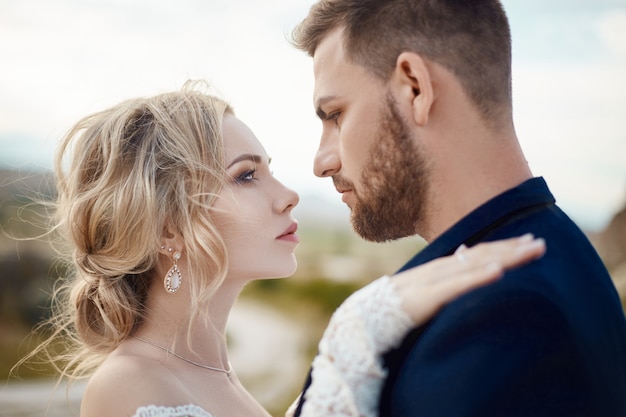 Couple in love hugs and kisses in fabulous mountains in nature. Girl in long white dress with bouquet of flowers in her hands, man in jacket. Wedding in nature, relationships and love