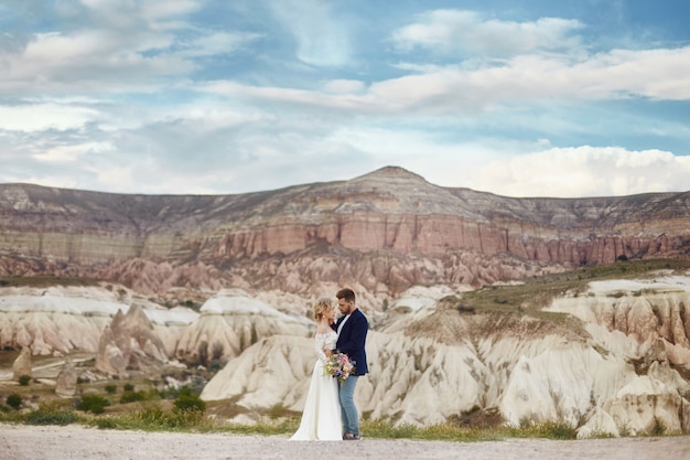 Couple in love hugs and kisses in fabulous mountains in nature. woman in long white dress with bouquet of flowers in her hands, man in jacket. Wedding in nature, relationships and love