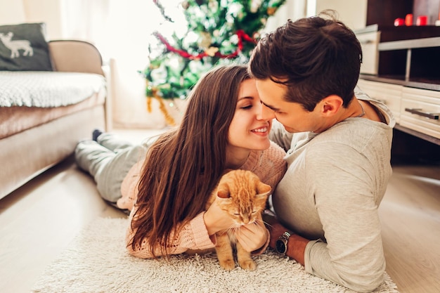 クリスマスツリーのそばに横たわって、家で猫と遊ぶ愛のカップル。幸せな男と女のキス