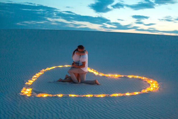 Couple in love romantic hugs in sand desert. Evening, romantic atmosphere, in sand burn candles in form of heart