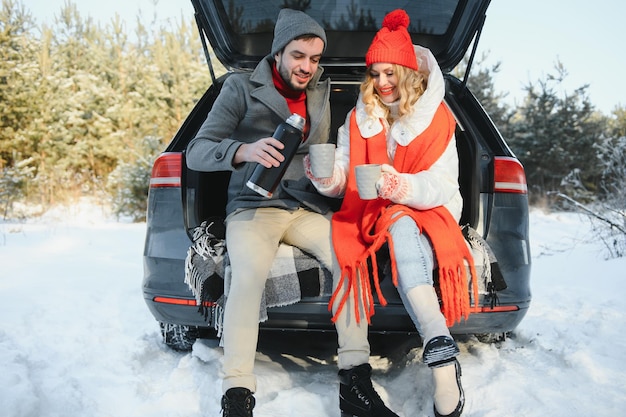 Couple in love sitting in car trunk drinking hot tea in snowy winter forest and chatting. People relaxing outdoors during road trip. Valentines day