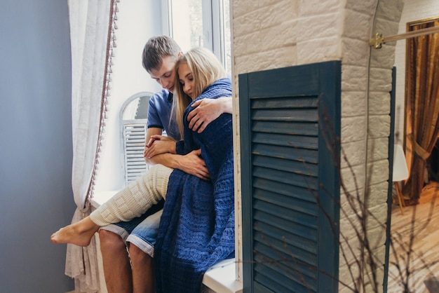 窓に家で座っている愛のカップル。新婚夫婦の優しい愛情のこもった抱擁。愛するカップルの楽しい朝の幸せな気分。女の子は彼氏のロマンチックな気持ちに寄り添います。バレンタイン・デー