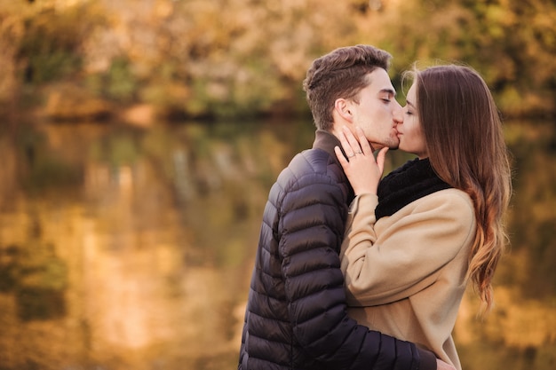 黄色の落ち葉で秋の公園で立っている愛のカップル。男と女が一緒に一日を楽しんでいます。唇で女の子にキスする少年