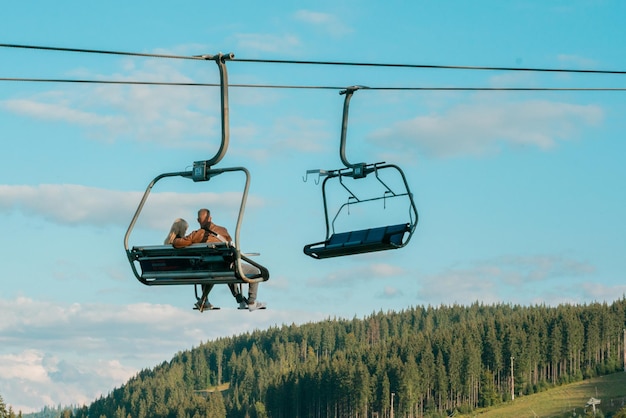 A couple in love in the summer time climb a mountain on a chair\
lift. wood. forest. recreation. leisure. climbs up. nature.\
romantic. elevator. family. national park