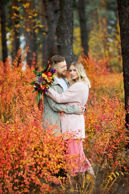 Couple in love walks through autumn forest. Hugs and kisses of men and woman, relationships and love. Young couple stands in yellow red grass, a bouquet of flowers in girl hand