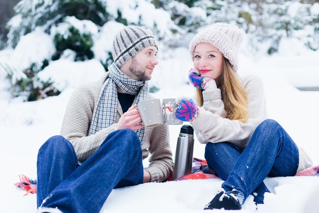 暖かい服を着た愛のカップルが雪の中で座って、温かい飲み物でカップを保持