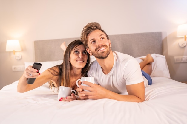 A couple in love watching TV on top of the hotel bed in pajamas