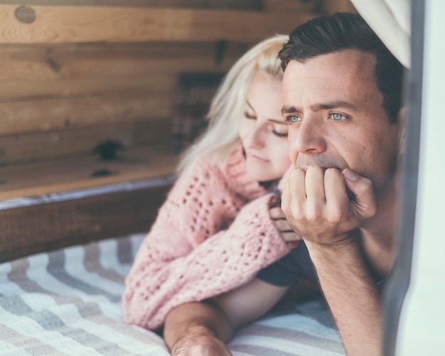 Couple in love with adult man looking ahead and thoughtful and blonde young girl hugged to him Different age relationship people boy and girl Love and problems Relax laying on bed inside cabin