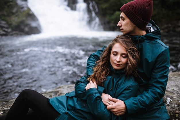 A couple of lovers in green raincoats, sitting on a rock, against the backdrop of a waterfall