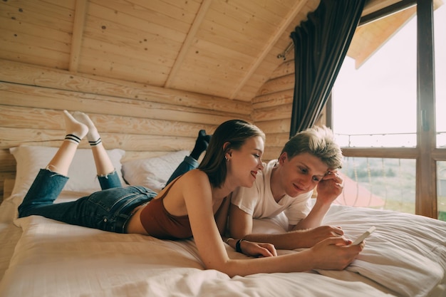 пара лежит на кровати в квартире с деревянным интерьером, смотрит на экран смартфона и улыбается