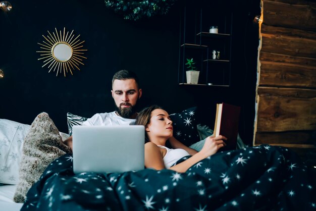 Couple lying in bed reading book and using laptop