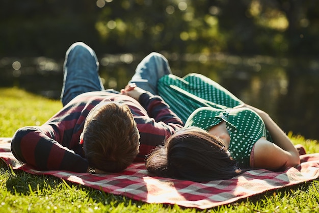 Couple lying and blanket for by lake at park with care love and bonding on grass in spring sunshine Man woman and outdoor together with connection holiday or relax by water for date in nature