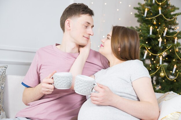 Coppia uomo e donna che bevono tè caffè seduti sul divano con decorazioni natalizie sullo sfondo.