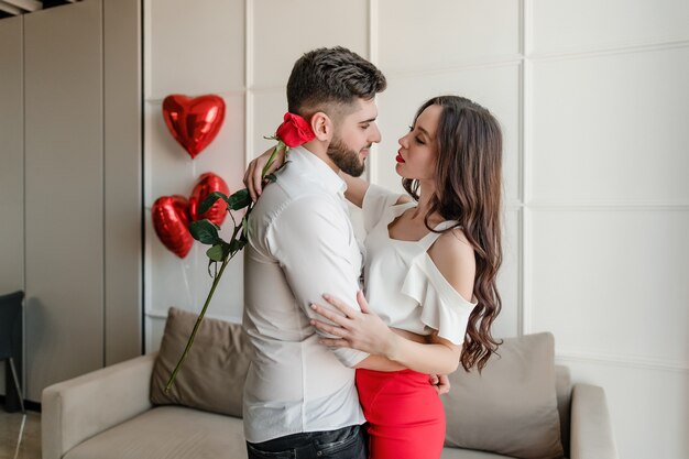 Couple man and woman in love with red rose and heart shaped balloons at home
