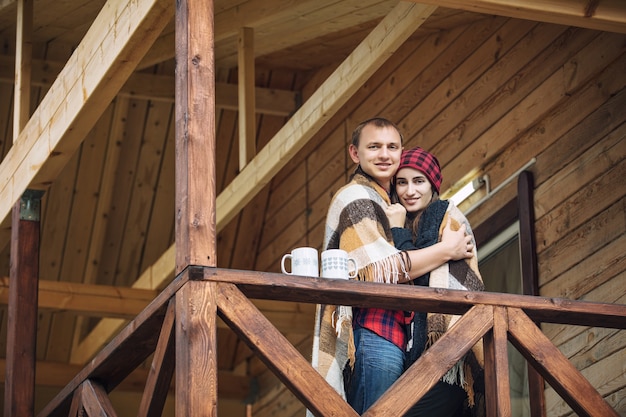 Coppia uomo e donna giovane bella felice sotto il portico di una casa in legno in natura con tazze di bevande calde